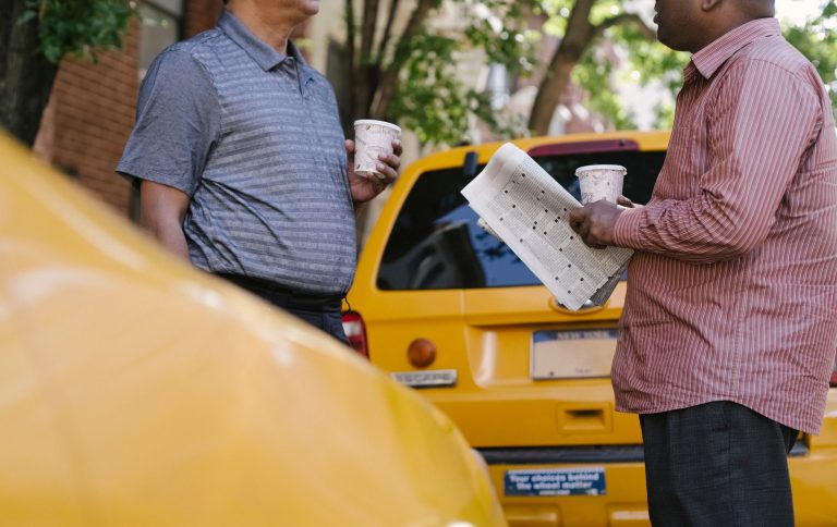 Crop anonymous multiracial taxi drivers with takeaway coffee and newspaper speaking near yellow cars in town — Pourquoi les c